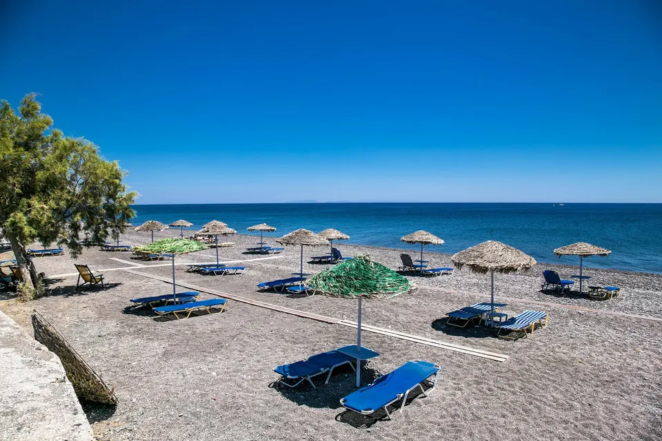 Sort strand med stråparasoller, blå solsenge og et tamarisktræ