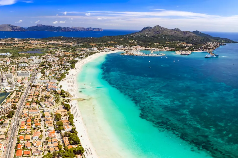 Luftfoto af Alcudia-bugten på Mallorca med lang hvid sandstrand, lavt turkist vand og Port d'Alcúdia