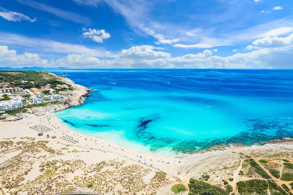 Luftfoto af Cala Mesquida med den brede sandstrand, klitterne og turkis vand under den lille bebyggelse