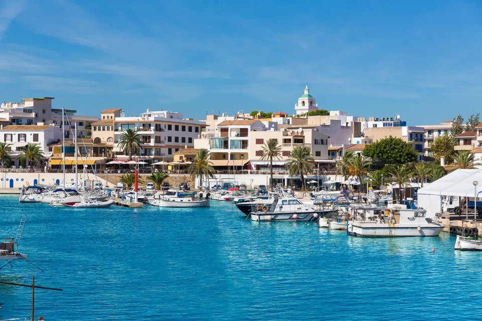 Havnen i Cala Ratjada med fiskerbåde, turkis vand og husene langs havnefronten under kirkekuplen