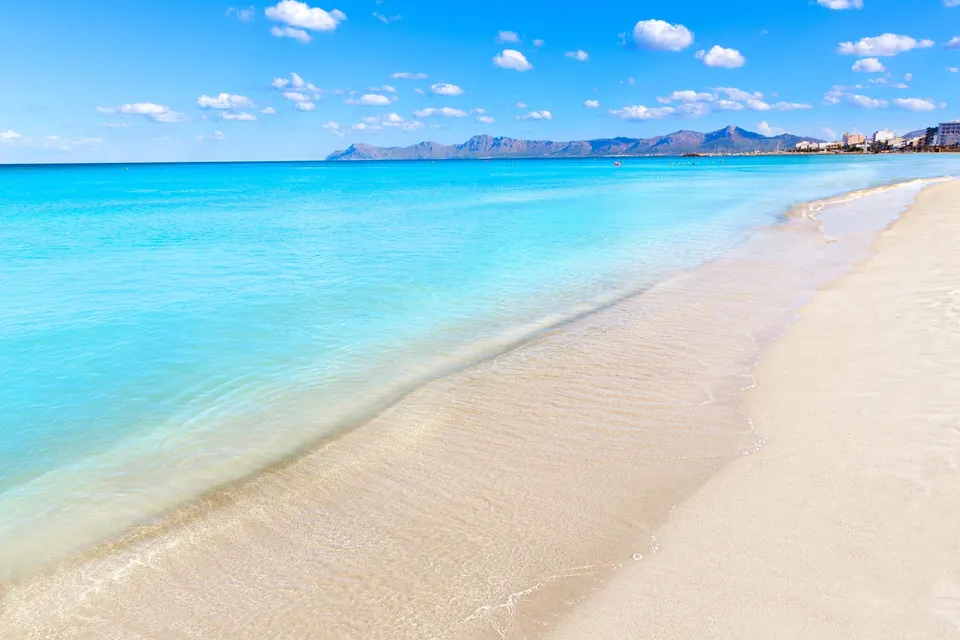 Lys sandstrand med lavt, turkis vand ved Can Picafort og bjergene om Alcudia-bugten i baggrunden