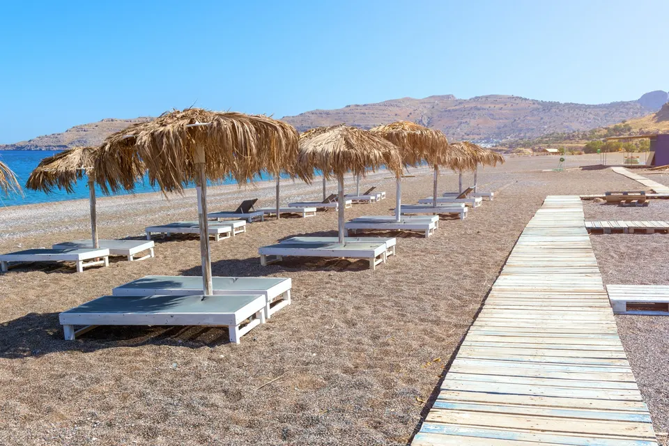 Solsenge under stråparasoller og en gangbro af træ på gruset strand ved Kalathos