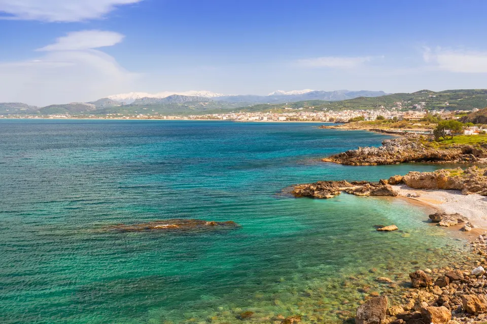 Kysten ved Kissamos på Kreta med turkist vand, klipper og byen under De Hvide Bjerge i baggrunden