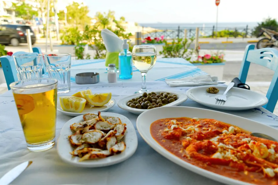 Mezedes, øl og hvidvin på et bord ved en promenade med havet bagved