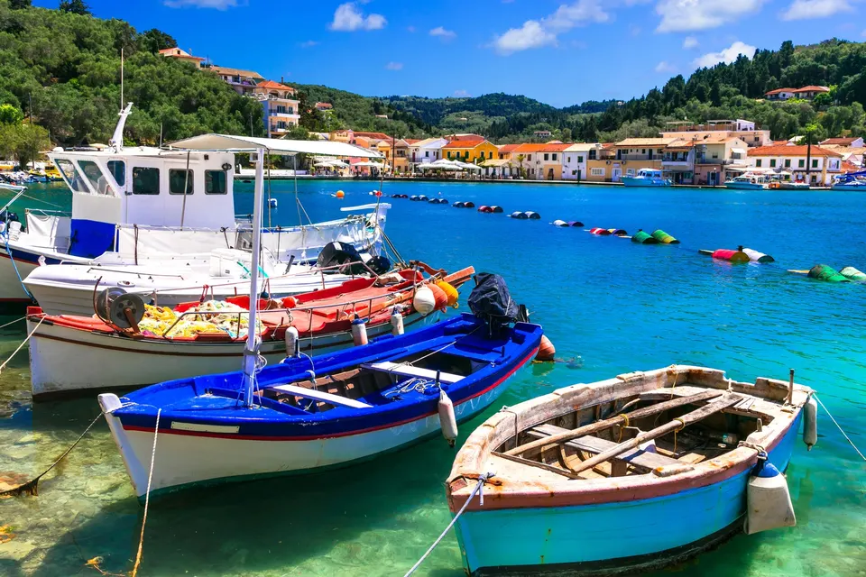 Farverige fiskerbåde i turkis vand i Lakka-bugten på Paxos med byen bagved