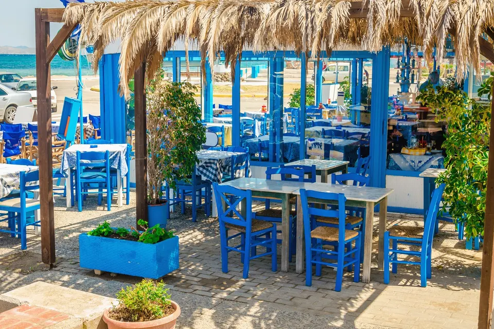 Taverna med blå stole under et tag af palmeblade med stranden lige bagved