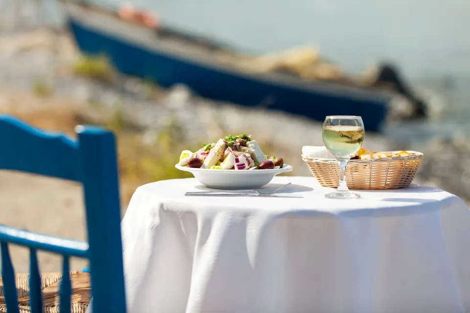 Bord med hvid dug, græsk salat og et glas hvidvin ved vandet