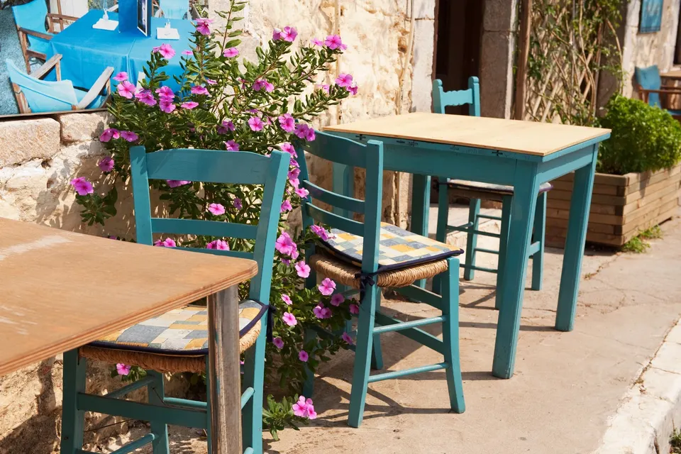 Turkise stole og borde på en terrasse ved en stenmur med lyserøde blomster